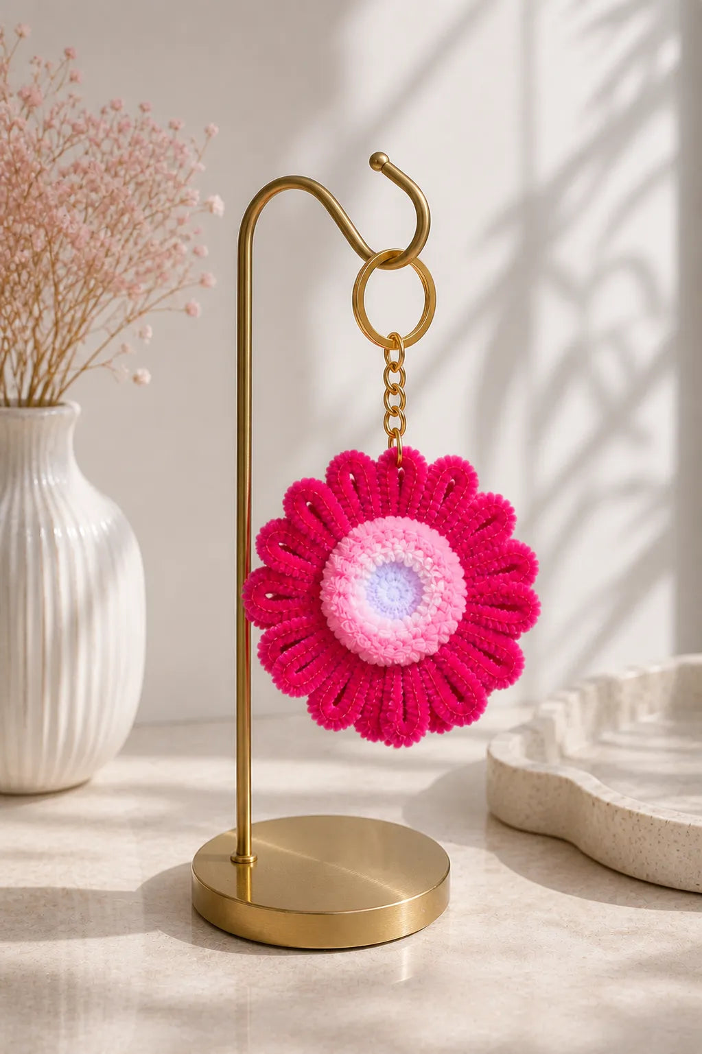 Pink crocheted flower keychain on a gold stand with a vase and decorative items in the background.